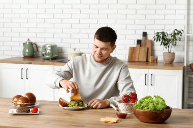 Genç adam, mutfakta lezzetli bir burger için çöreğe marul koyuyor.