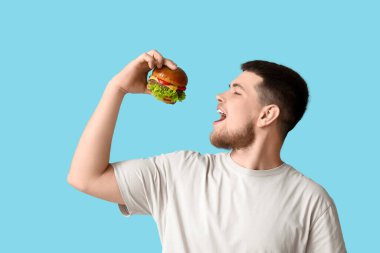Genç adam mavi arka planda lezzetli bir burger yiyor.
