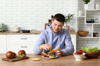 Genç adam mutfakta lezzetli hamburgerin üstüne peynir koyuyor.