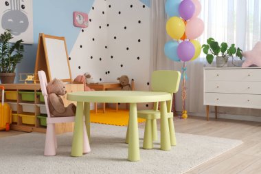Anaokulundaki modern oyun odasının içinde raf üniteleri, çekmeceler ve sandalyeli bir masa var.