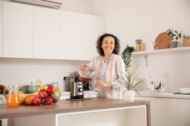 Olgun bir kadın mutfakta blenderdan cama meyve suyu dolduruyor.