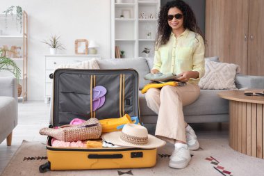 Genç Afro-Amerikalı kadın otel odasındaki kanepede seyahat aksesuarlarıyla bavulunu boşaltıyor.