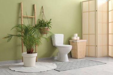 Tuvaletin içinde klozet, çöp tenekesi, katlanan ekran ve yeşil duvarın yanında bitki var.