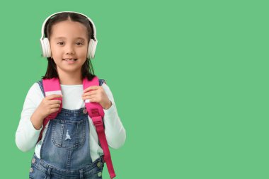 Yeşil arka planda sırt çantalı, kulaklıklı şirin, mutlu bir Asyalı kız. Okul yılı sonu.
