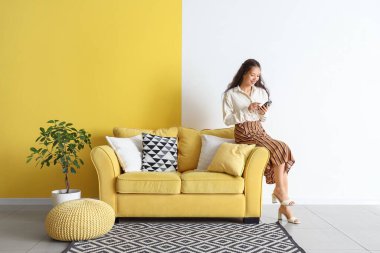 Duvarın yanındaki sarı koltukta cep telefonu kullanan genç Asyalı kadın.