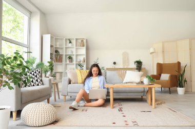 Genç Asyalı kadın evdeki gri kanepenin yanındaki halıda dizüstü bilgisayar kullanıyor.