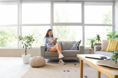 Genç Asyalı kadın evdeki gri koltukta tablet bilgisayar kullanıyor.