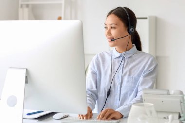 Kulaklıklı genç bayan resepsiyonist ofiste modern bilgisayarla işyerinde oturuyor.