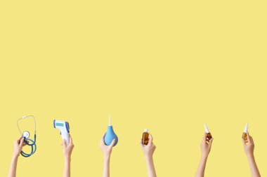 Sarı arka planda steteskop, termometre, lavman ve burun damlası olan kadın eller.