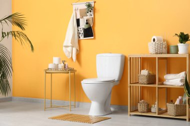 Tuvaletin içinde beyaz klozet, sehpa ve turuncu duvarın yanında raf ünitesi var.