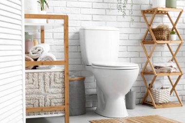 Beyaz tuğla duvarın yanında seramik klozet, ev bitkisi ve raf ünitesi olan güzel bir tuvalet.