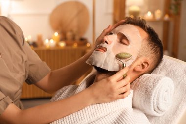 Maskeli yakışıklı genç adam güzellik salonunda yüz masajı yaptırıyor.