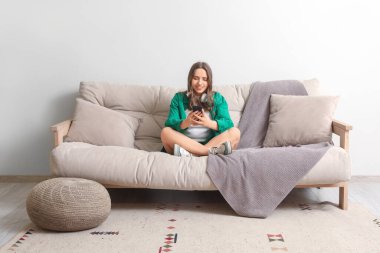 Kulaklıklı güzel bir kadın evdeki gri koltukta cep telefonu kullanıyor.