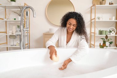Evinde banyo süngeri ve sabun köpüğüyle bornoz giymiş güzel, mutlu, Afro-Amerikan bir kadın.