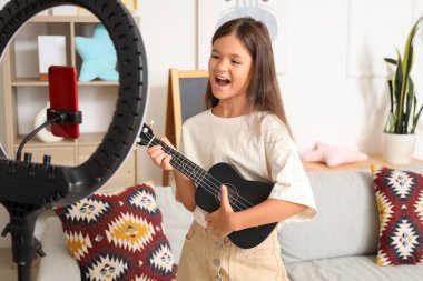 Evde mikrofon ve ukulele ile şarkı söyleyen küçük bir kız.