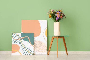 Yeşil duvarın yanındaki sehpanın üzerinde yabani çiçekler olan resimler ve vazolar.