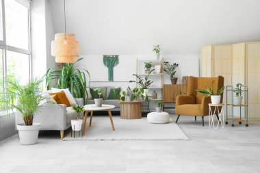 Yeşil bitkilerle, kanepelerle ve masalarla aydınlık oturma odasının içinde.