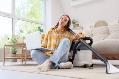 Dizüstü bilgisayarı ve elektrikli süpürgesi olan yorgun genç bir kadın evdeki halıda oturuyor.