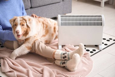 Battaniyeli genç bir kadın ve Avustralyalı çoban köpeği ısıtıcının yanında oturuyorlar.