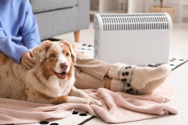 Battaniyeli genç bir kadın ve Avustralyalı çoban köpeği ısıtıcının yanında oturuyorlar.