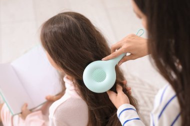 Genç anne, banyoda yerde oturmuş kitap okuyan küçük kızının saçını tarıyor.