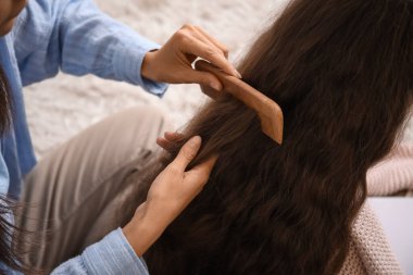 Genç anne, evde yerde otururken kızının saçlarını tarıyor.