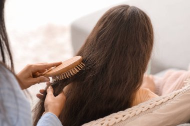 Genç anne, evdeki kanepede otururken küçük kızının saçını tarıyor.