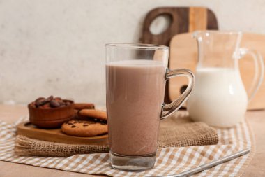 Bir bardak çikolatalı süt, kakao çekirdekleri ve bej masada lezzetli kurabiyeler.