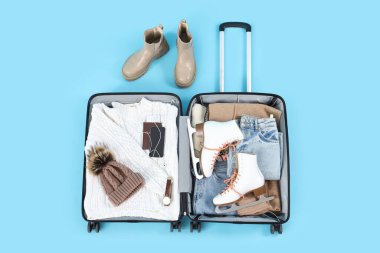 Mavi arka planda kış giysileri ve buz patenleriyle dolu bavulu aç.