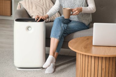 Elinde çay olan genç bir kadın evde hava temizleyiciyi açıyor.