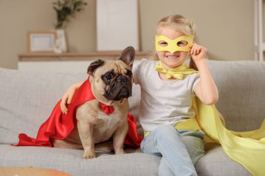 Evdeki kanepede oturan süper kahraman kostümlü Fransız bulldog 'u olan şirin mutlu küçük kız.
