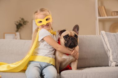 Evdeki kanepede oturan süper kahraman kostümlü Fransız bulldog 'u olan şirin mutlu küçük kız.