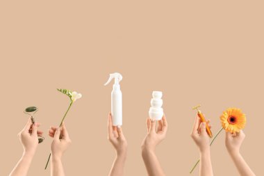 Kozmetik ürünleri şişeleri, yüz masajı aletleri ve bej arka planda çiçekler olan birçok kadın eli.
