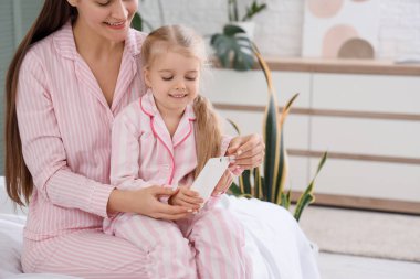 Sevimli küçük mutlu kız ve annesi evde yatağında krema şişesiyle pijama giymiş.