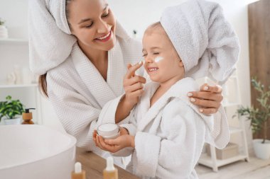 Sevimli küçük mutlu kız ve bornozlu annesi evde krem sürüyor.