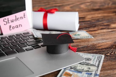 Küçük mezuniyet şapkalı modern dizüstü bilgisayar, ÖĞRETMENE kredisi, para ve diploma ahşap masada, yakın plan