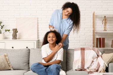 Mutlu Afro-Amerikalı anne, tatlı kızının kıvırcık saçlarını evdeki kanepede spreyliyor.