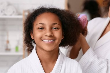 Afro-Amerikalı anne mutlu kızının kıvırcık saçlarını banyoda tarıyor.