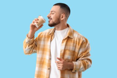 Genç adam mavi arka planda kağıt bardaktan kahve içiyor.