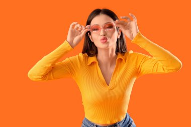 Güneş gözlüklü genç kadın turuncu arka planda öpüşüyor.