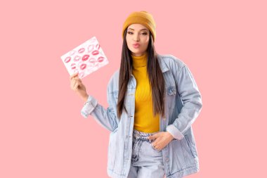 Pembe arka planda ruj izleri olan öpücük ve kağıt tutan genç bir kadın.