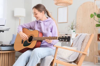 Evdeki koltukta gitar çalan genç bir kadın.