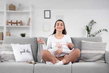 Evdeki kanepede meditasyon yaparken bebeğini emziren genç bir kadın.