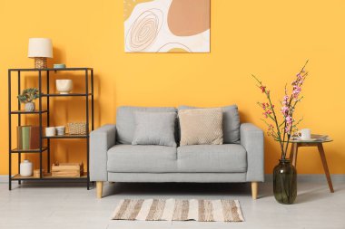 Konforlu kanepe, raf ünitesi ve sarı duvarın yanında çiçek açan ağaç dalları.