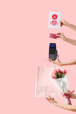 Kadın eller ödeme terminalini kağıt torba, kartpostal ve pembe arka planda çiçekler ile tutuyor. Uluslararası Kadınlar Günü için Alışveriş