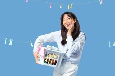 Çamaşır sepeti ve mavi arka planda çamaşır mandallarıyla mutlu bir Asyalı kadın.