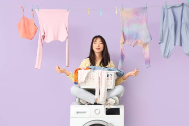 Çamaşır, sepet ve çamaşır mandallarıyla meditasyon yapan genç bir Asyalı kadın mor arka planda çamaşır makinesinde oturuyor.