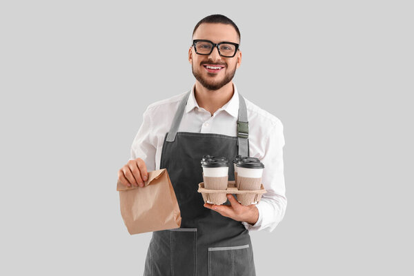 Молодой бариста с бумажными чашками кофе и едой на сером фоне