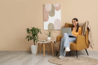 Genç bir kadın dizüstü bilgisayar kullanıyor ve oturma odasının içindeki bej duvarın yanındaki koltukta oturuyor.