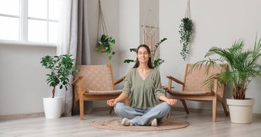 Genç bir kadın evde yerde oturmuş meditasyon yapıyor.